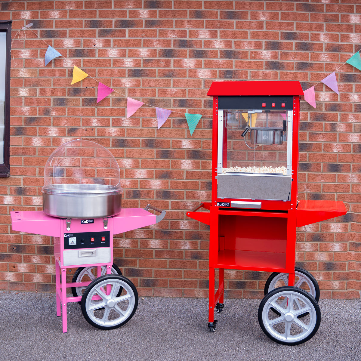 Een roze suikerspinmachine en een rode popcornmachine staan naast elkaar op een stenen vloer, met kleurrijke vlaggetjes op de achtergrond