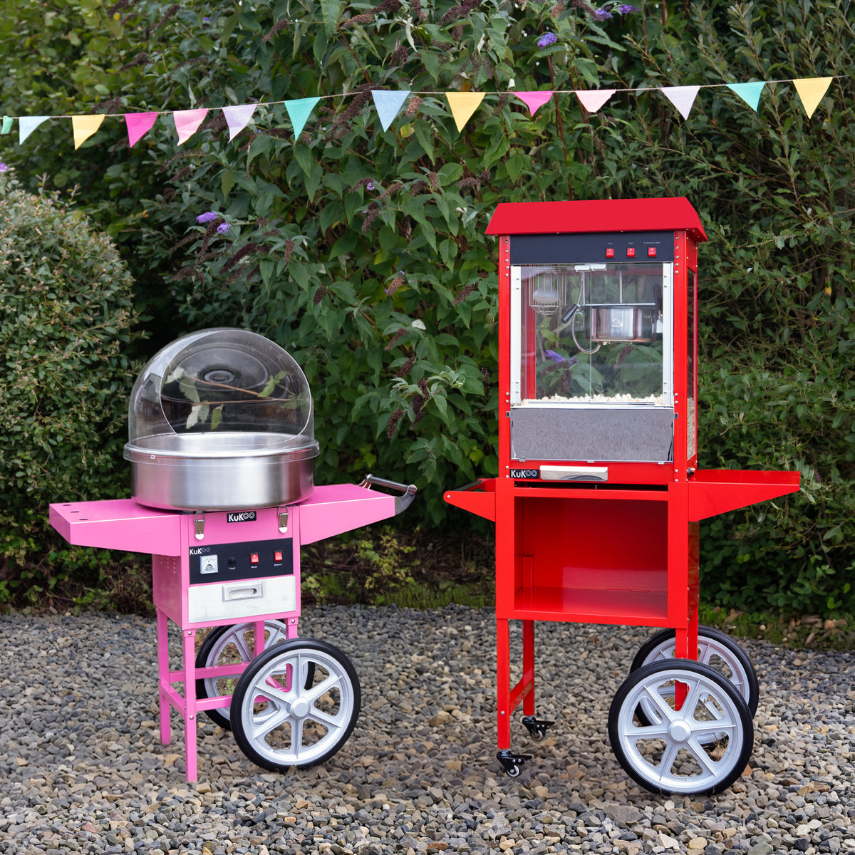 Roze suikerspinmachine en rode popcornmachine op wielen, geplaatst op een grindpad met groene planten en vlaggen op de achtergrond