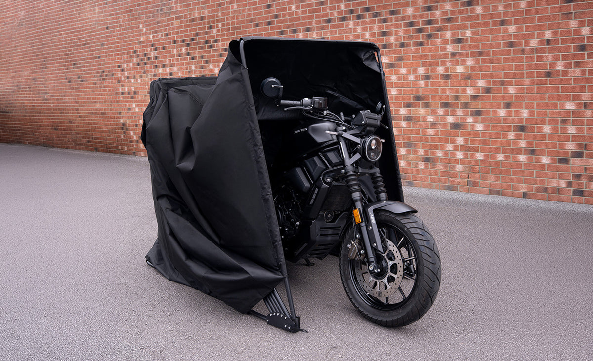 Zwarte motorfiets gedeeltelijk bedekt met een waterdichte hoes, geplaatst op een grijze ondergrond tegen een bakstenen muur