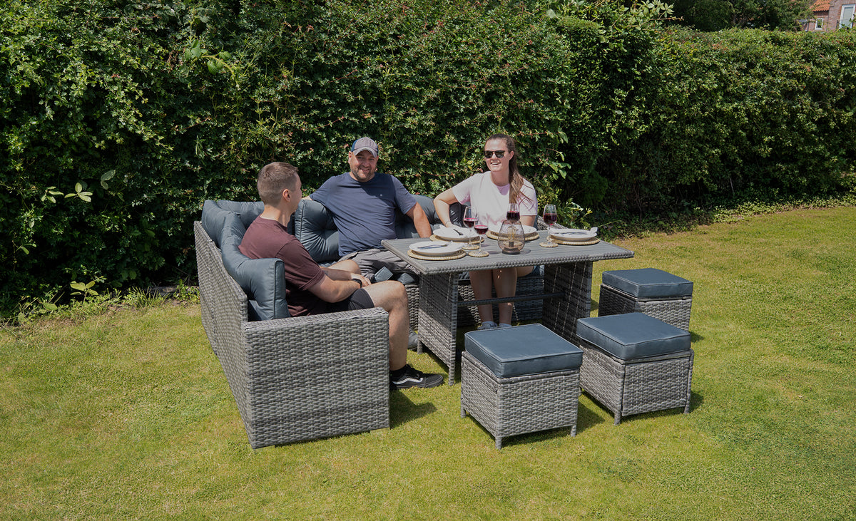 Drie mensen zitten aan een moderne, grijze wicker tuinset met kussens, genietend van een maaltijd in een groene tuin