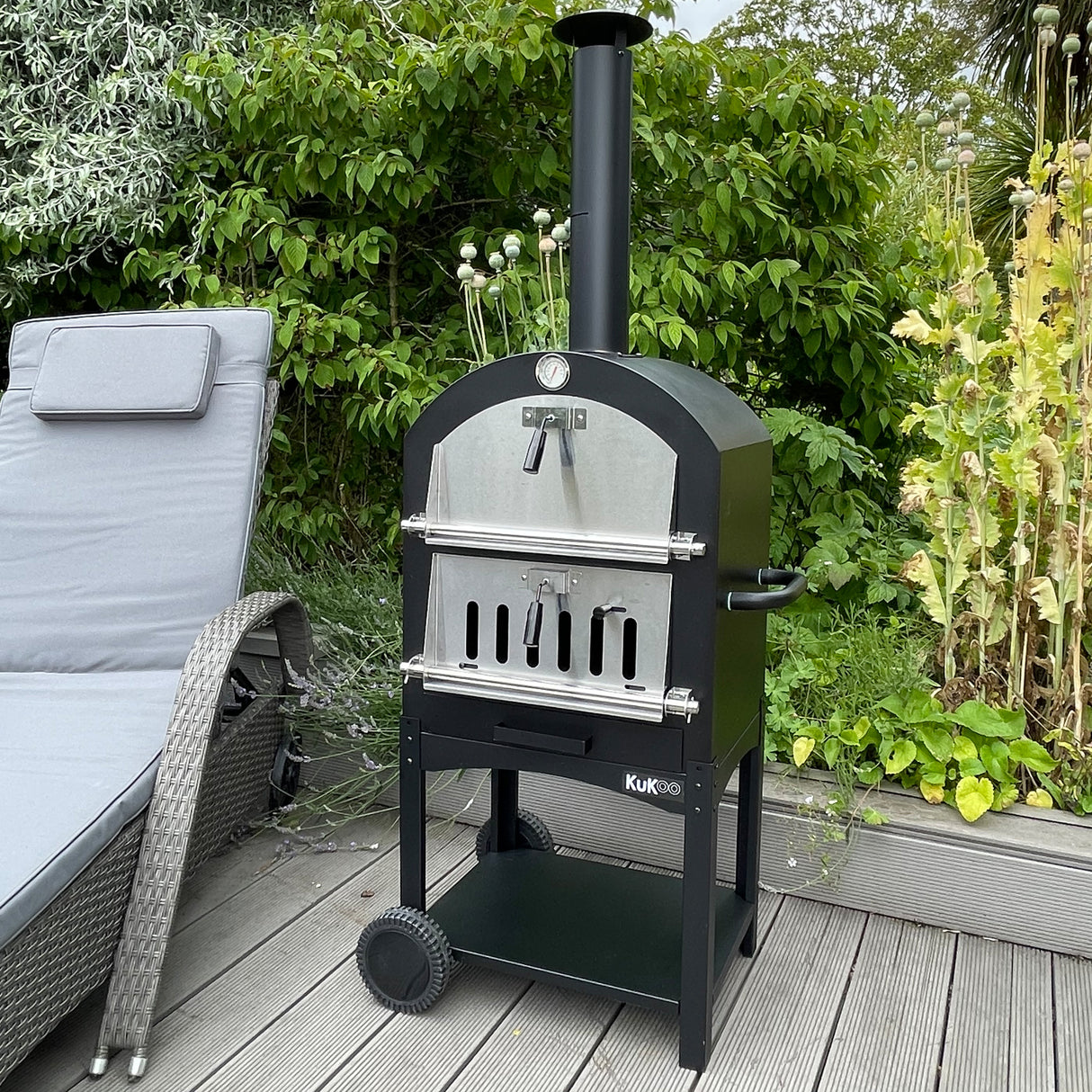 Zwarte KuKoo buiten pizzaoven met roestvrijstalen deur, op een houten terras omringd door groene planten en een comfortabele ligstoel