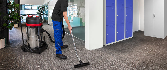 Een man in blauwe werkbroek stofzuigt een moderne kantoorruimte met een krachtige stofzuiger, omringd door groene planten en blauwe lockers