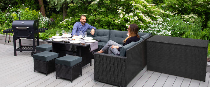 Moderne tuinset met zwart wicker, comfortabele kussens, een glazen tafel en een barbecue, omringd door groene planten en bloemen